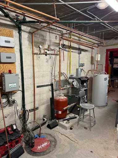 Basement utility room with pipes, boiler, water heater, tools, and cleaning equipment on a concrete floor