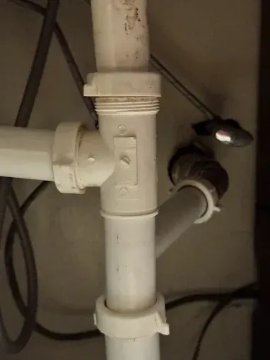 White PVC plumbing pipes and joints under a sink, with a small metal light fixture in the background