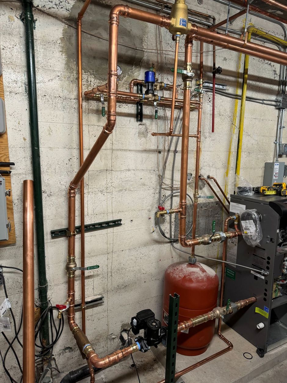 Basement utility room with copper pipes, a red expansion tank, and boiler equipment against a cinderblock wall
