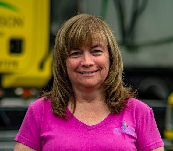 A woman in a pink shirt is smiling in front of a yellow truck.