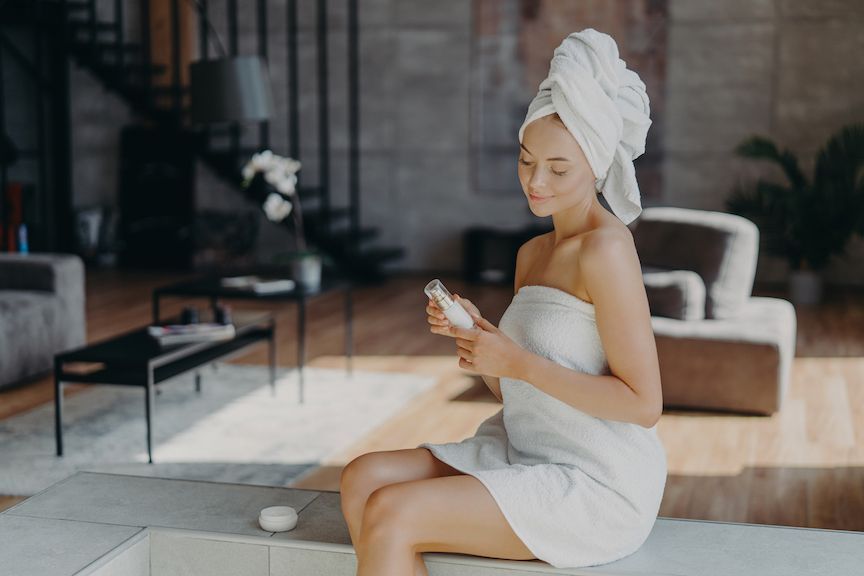 A woman wrapped in a towel is sitting on a ledge looking at a bottle of lotion.