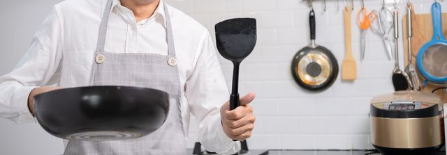 A man in an apron is holding a wok and spatula in a kitchen.