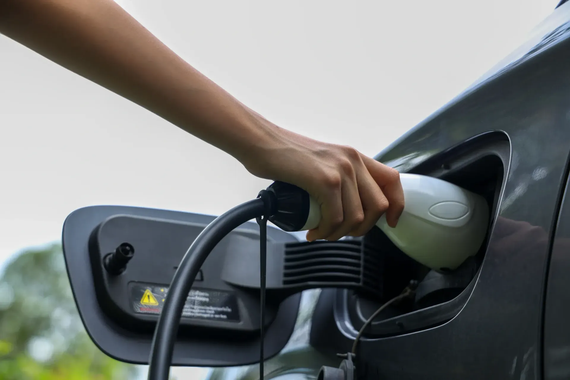 Hand plugging a charging cable into an electric vehicle's port. The car is gray, and the background is a blurred sky.