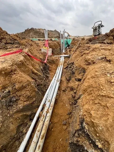 Trench in construction site with white conduits laid, dirt walls, cloudy sky.