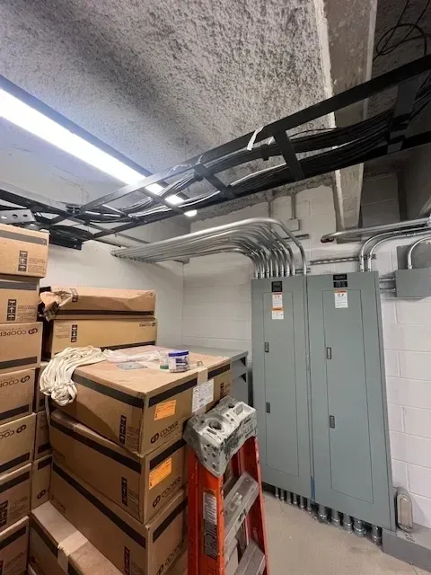 Electrical room with conduit, electrical panels, boxes, and a ladder. Grey panels and ceiling contrast with the white walls.