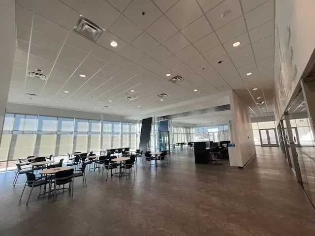 A large, open room with tables and chairs. Floor-to-ceiling windows on the left. Computers and glass-walled offices are visible in the background.