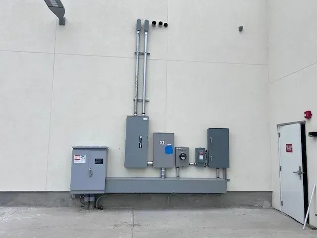 Electrical equipment mounted on a light-colored wall, including conduits, breaker boxes, and a gray trough. A white door is on the right.