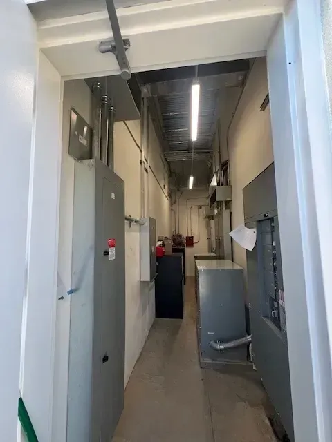 Narrow utility room with electrical equipment lining the walls. A long hallway leads to the back, lit by overhead fluorescent lights.