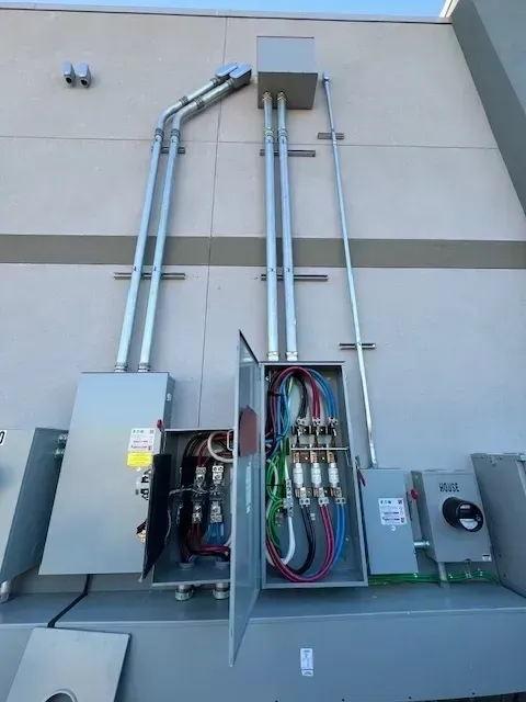 Exterior electrical panel on a building with conduits, wiring, and a meter. Gray and silver components against a neutral-colored wall.