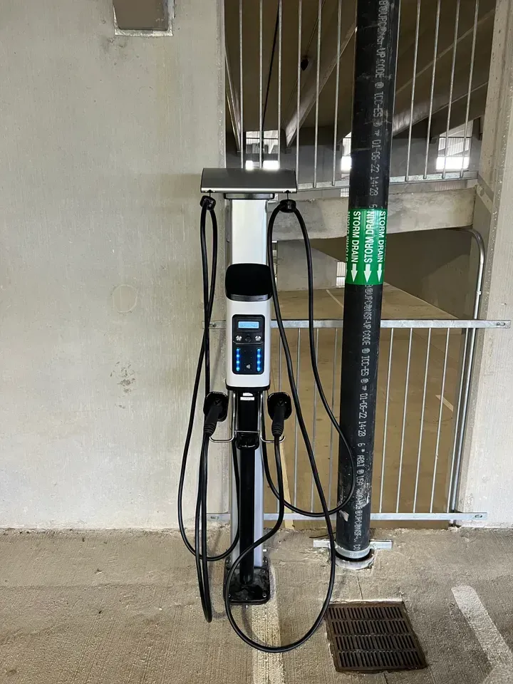 Electric vehicle charging station with two charging cables, mounted on a white pole next to a black pipe and concrete structure.