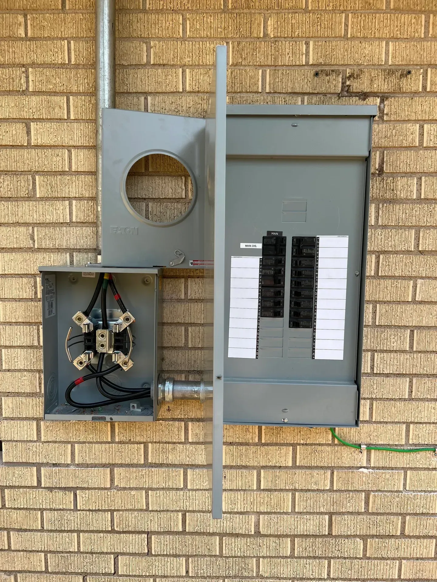 Electrical panel box and meter base mounted on a brick wall, with conduit and wiring exposed.