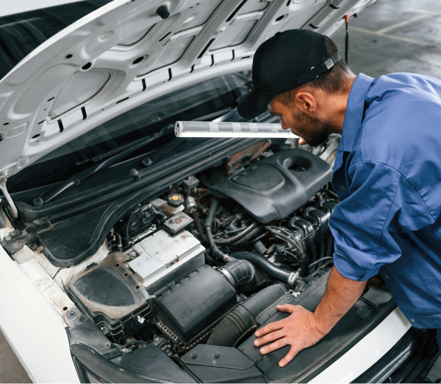 Mechanic inspecting a car engine, holding a light, in a well-lit garage, wearing blue work clothes and a cap | Ken's Import Service