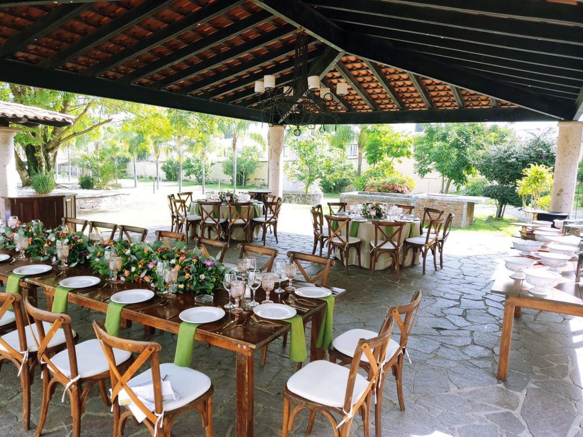 Mesas de recepción al aire libre dispuestas para una celebración, bajo un patio cubierto con detalles verdes.