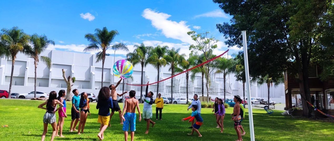Personas jugando con un globo de colores en un campo de hierba con palmeras y un cielo azul.