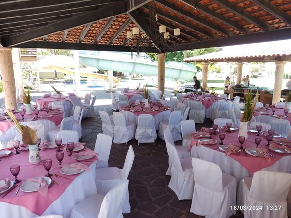 Mesas preparadas para una fiesta, con manteles rosas, fundas blancas para las sillas y cristalería morada bajo una pérgola.