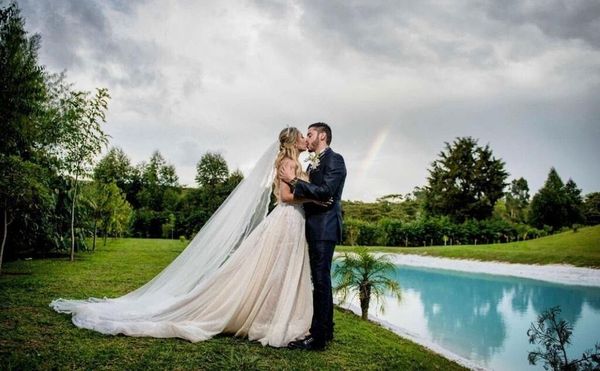 Novios besándose cerca de un lago azul claro, con árboles y cielo nublado. La novia lleva velo y vestido.