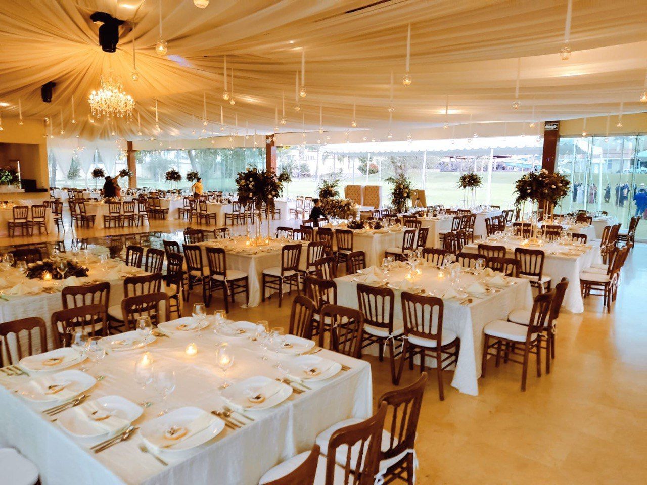 Salón de recepción formal preparado para una boda; mantelería blanca, sillas de madera y centros de mesa florales.