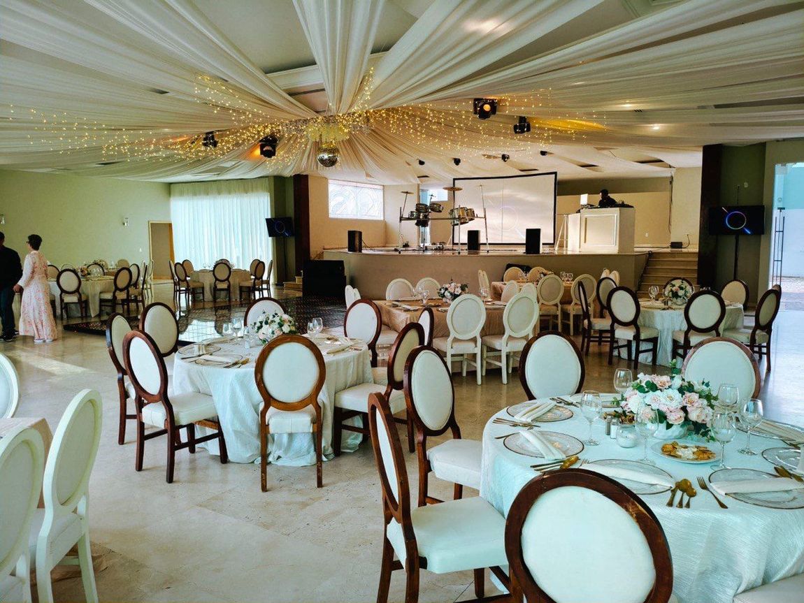 Elegante salón de banquetes con mesas redondas, sillas blancas y techo decorado con guirnaldas de luces.