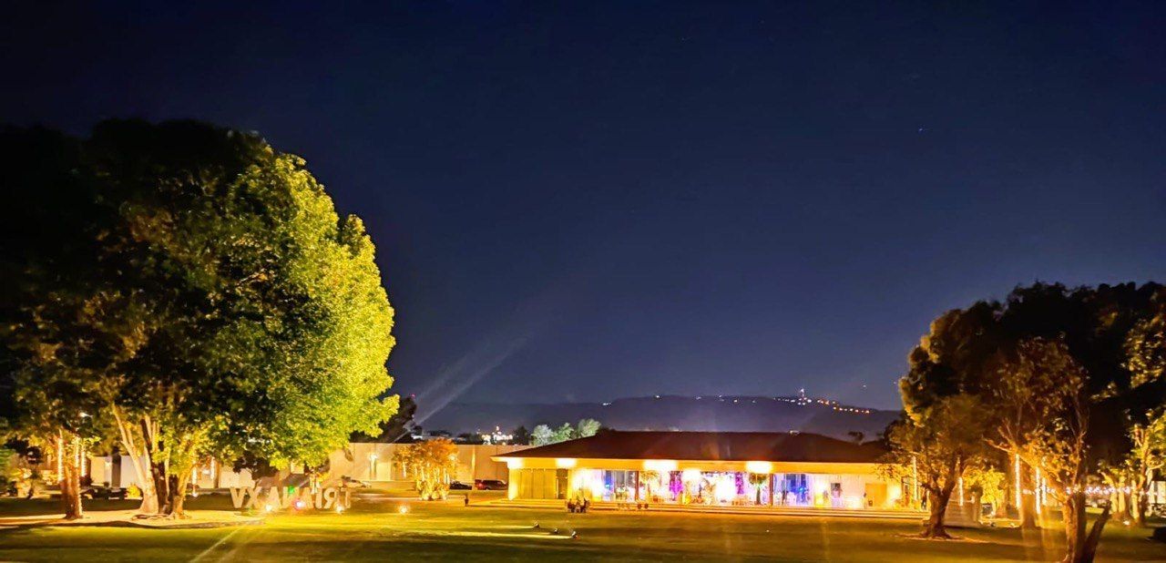 Vista nocturna de un edificio con las luces encendidas, flanqueado por árboles iluminados, bajo un cielo oscuro.