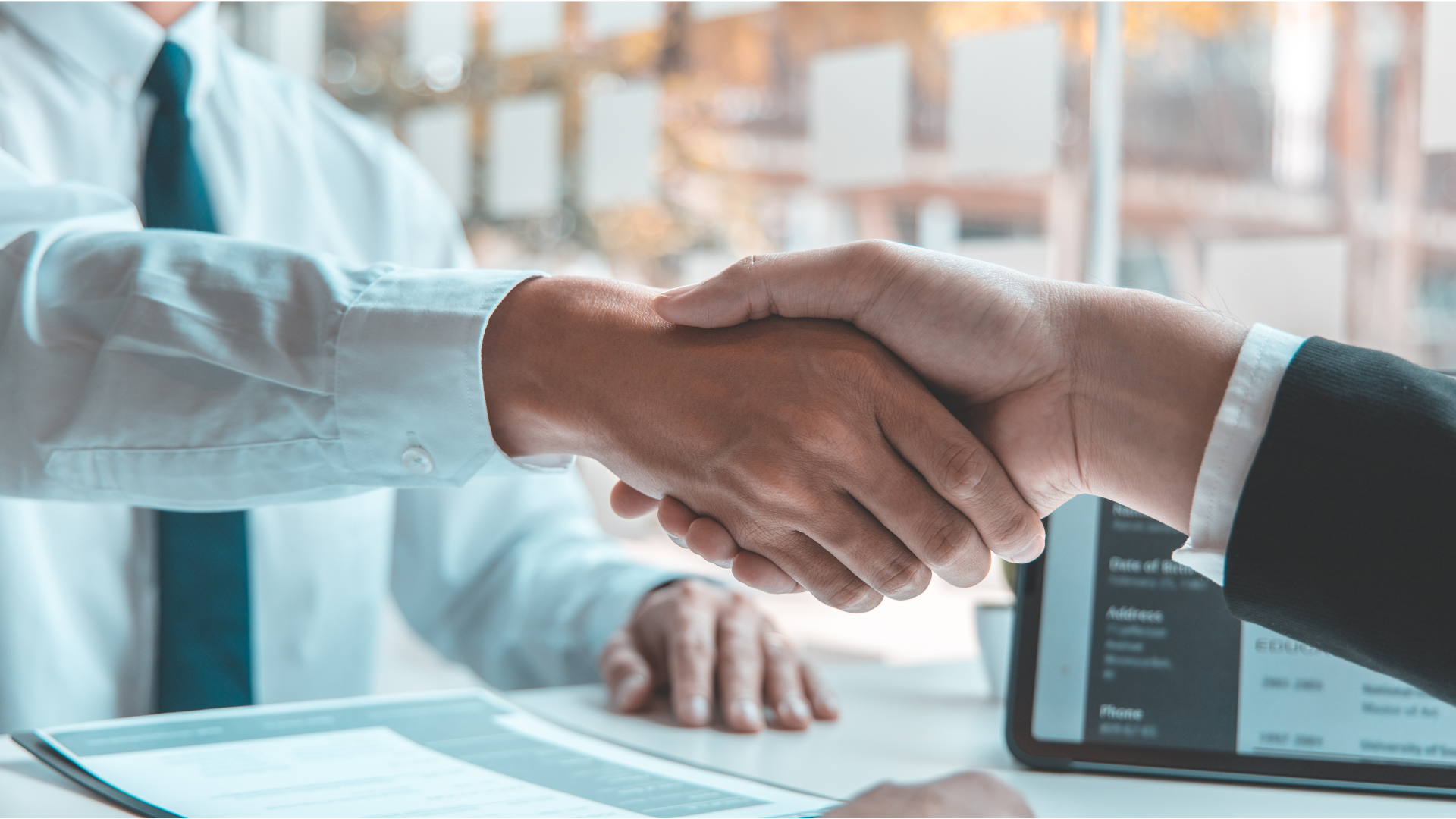 Two men shaking hands over a table. One in a suit jacket, the other in a shirt and tie example of human resources interview.