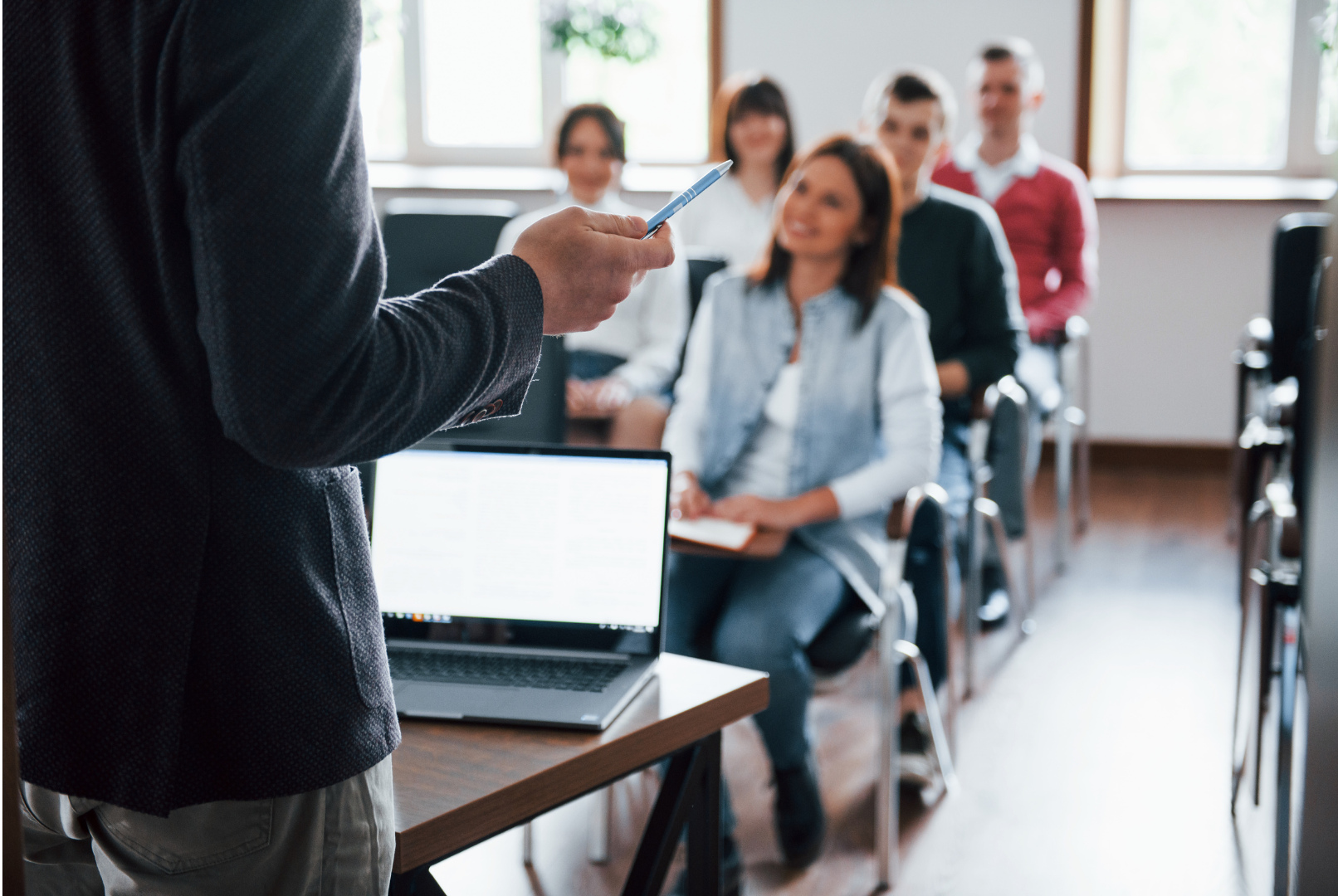 HR Advisory lecture points with pen at audience in a classroom; students seated, listening intently.