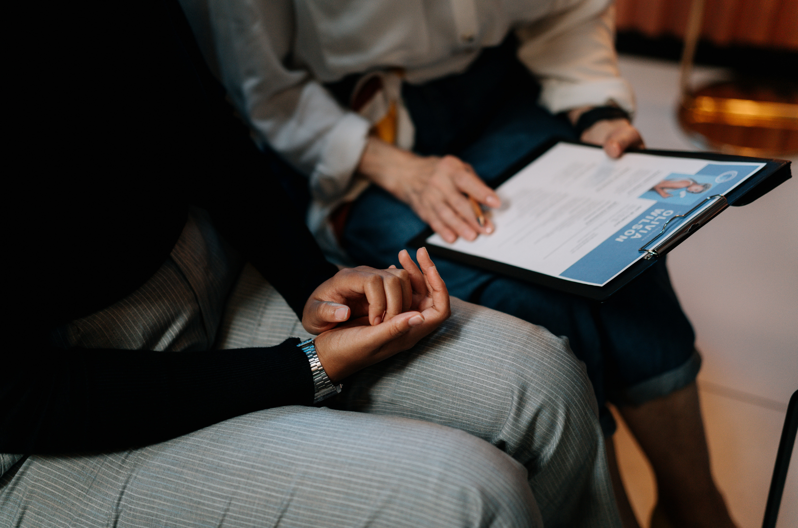 Fractional HR Person reviewing resume on clipboard; hands clasped, other person seated.