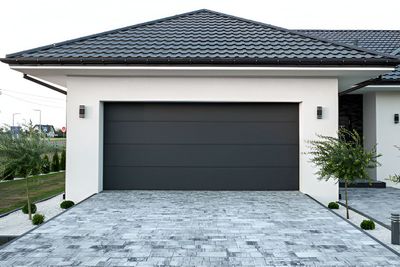 Gray garage door on a white building with a gray tiled driveway. Small bushes line the driveway.