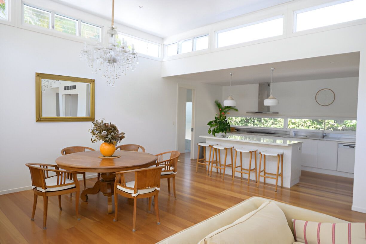 Open-plan dining and kitchen area with wooden floors and white walls. A round table is set for six, and a kitchen island has stools.