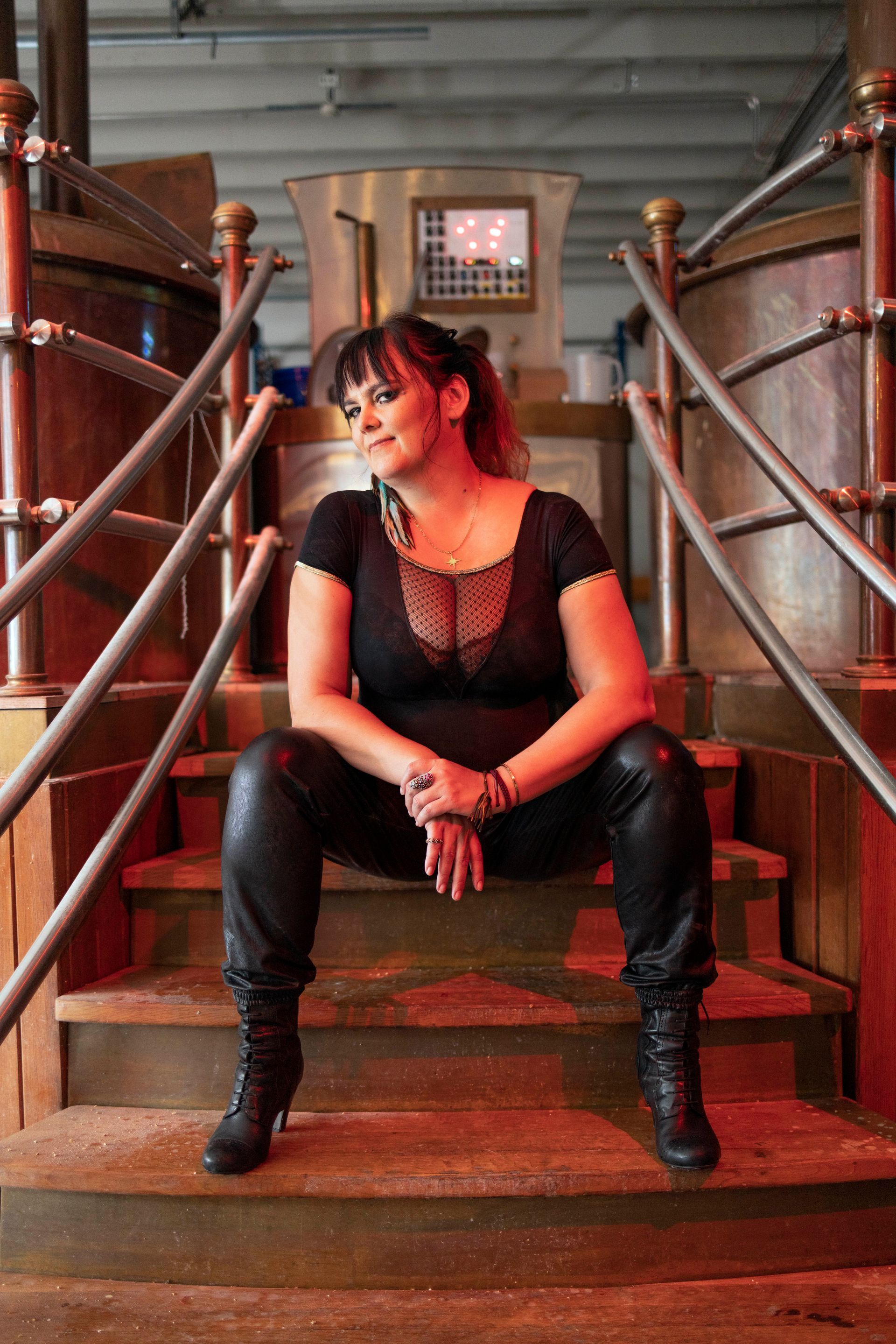 Woman in black outfit sits on brewery stairs, smiling, arms resting on knees.