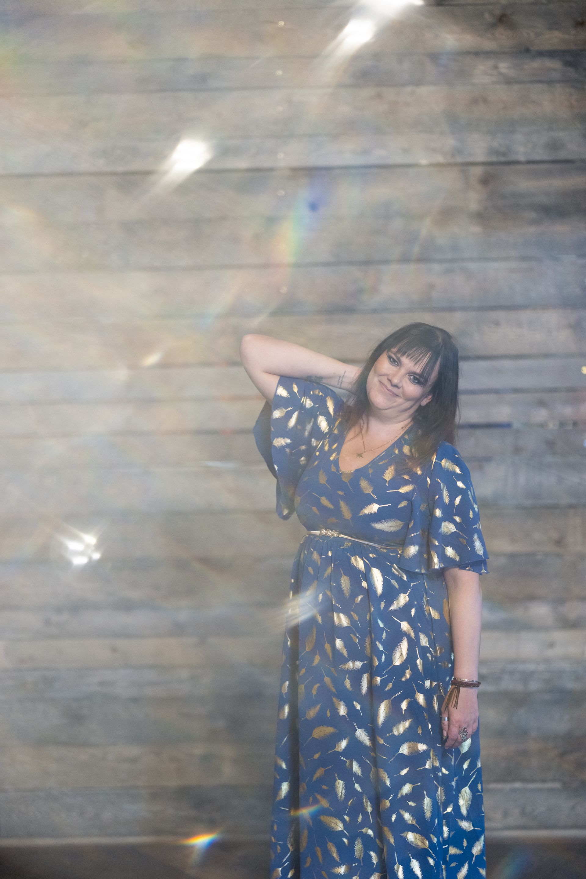 Woman in blue dress with gold leaf print, hand in hair, stands against wood panel wall.