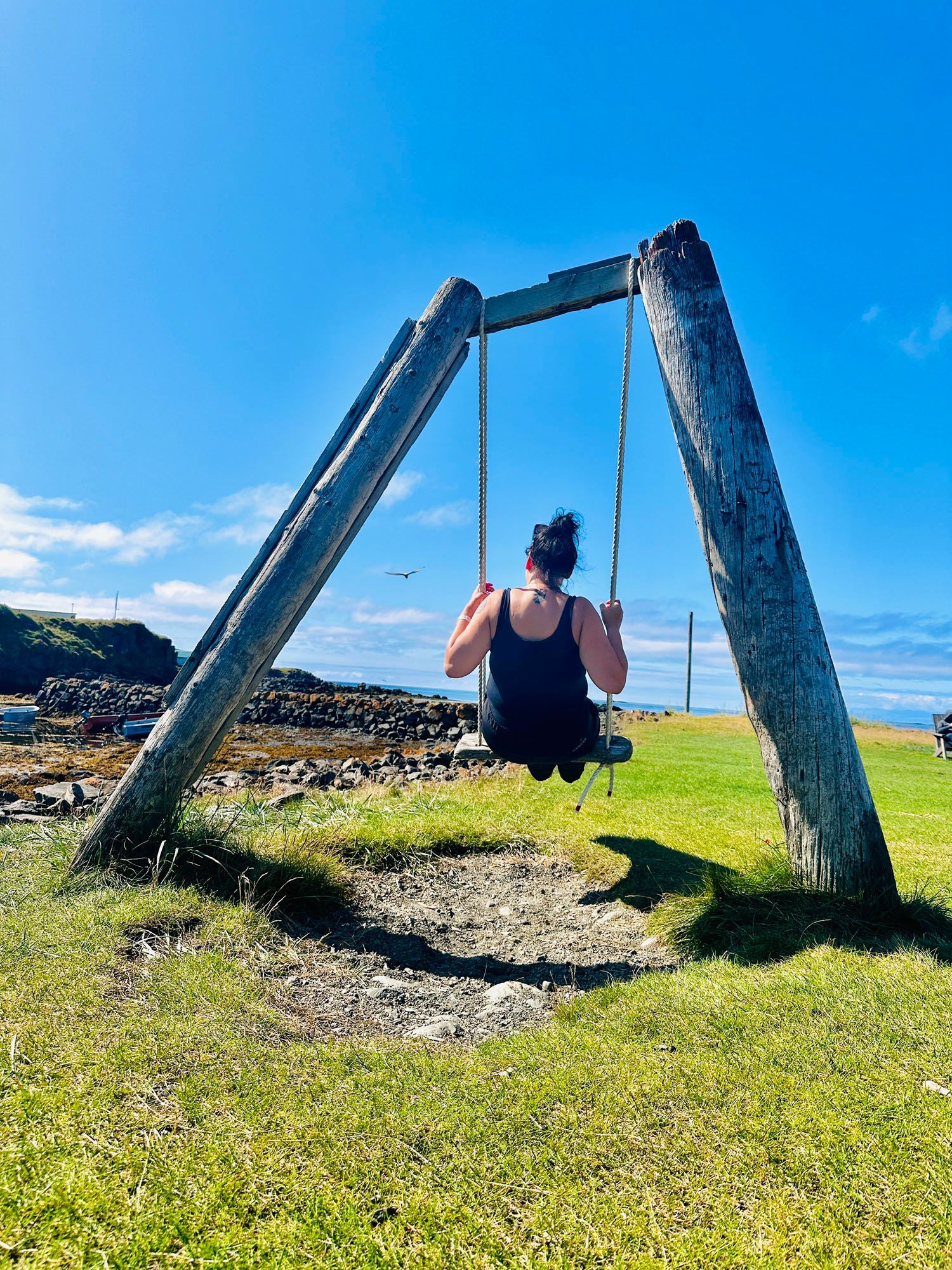 Woman on a swing set made of weathered wood, blue sky and ocean in the background.