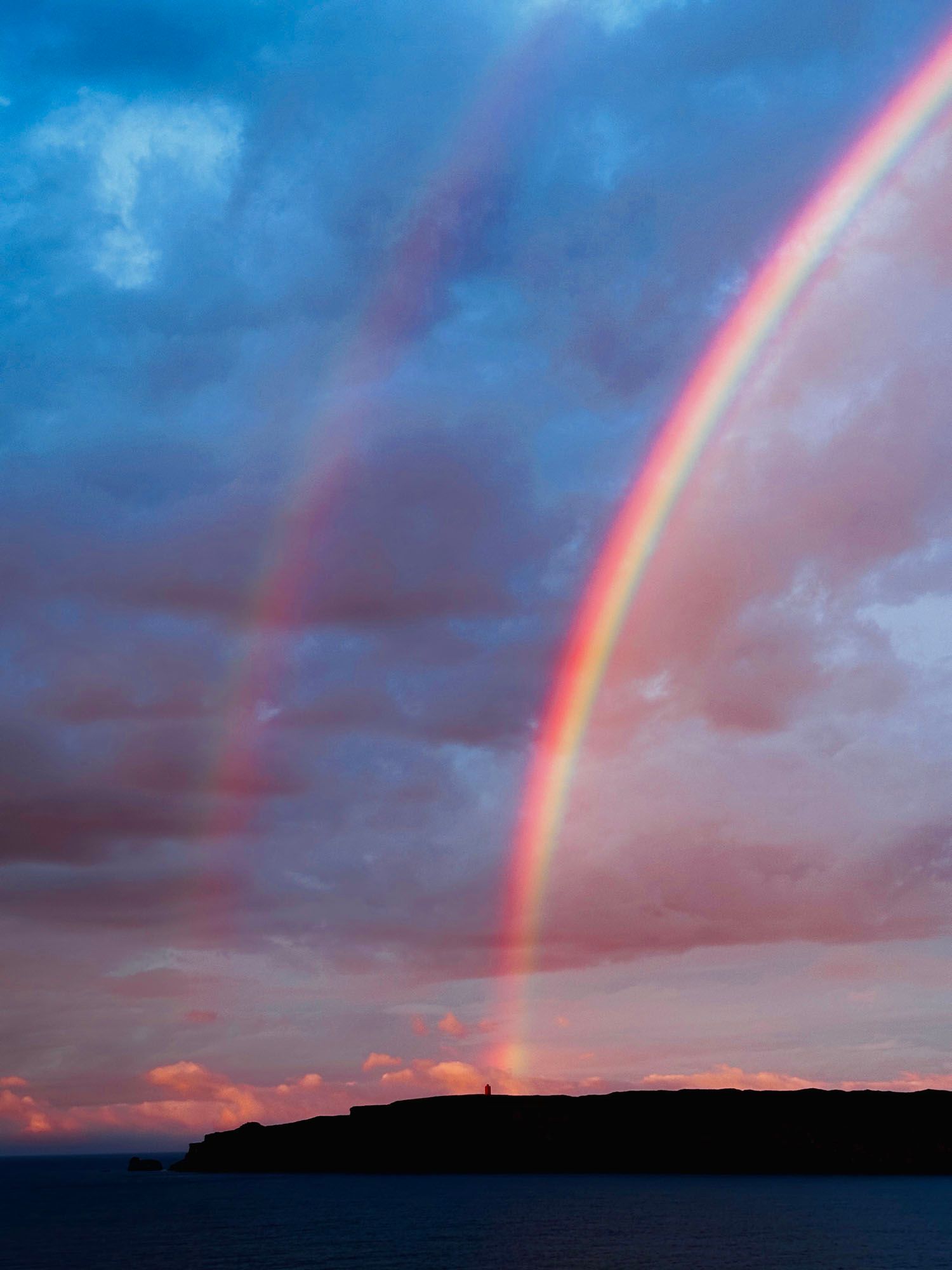 Double rainbow arches over a dark silhouette of a coastal landmass against a colorful, cloudy sky.