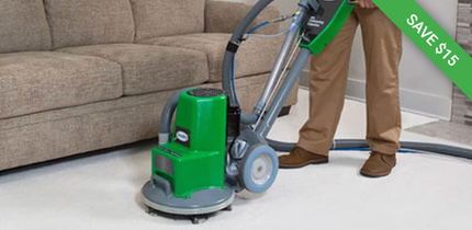 A person uses a green industrial floor cleaner on a white floor next to a beige sofa. A green tag reads 