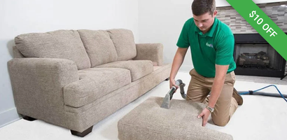 A professional in a green polo shirt cleans a sofa cushion with a handheld vacuum tool in a living room.