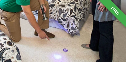 A person in a green shirt uses a UV light to inspect a carpeted floor in a bedroom, pointing out a stain to another person.