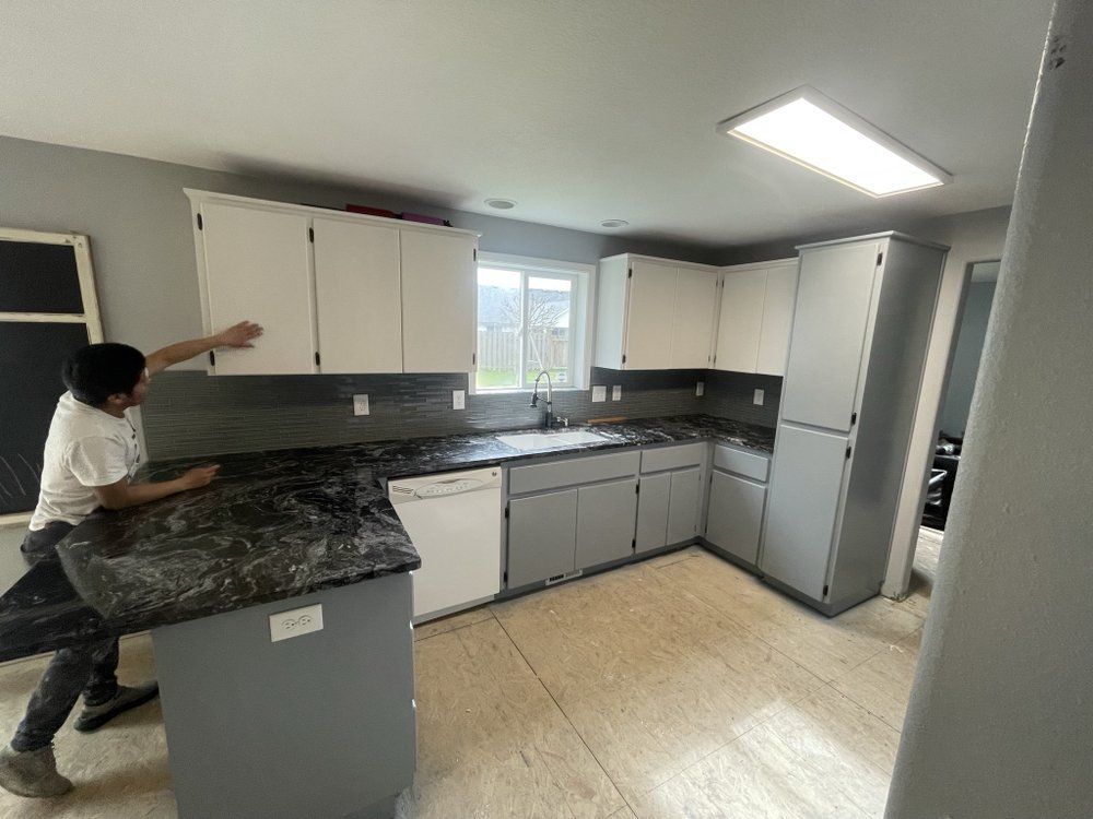 A man is standing in a kitchen with gray cabinets and a black counter top.