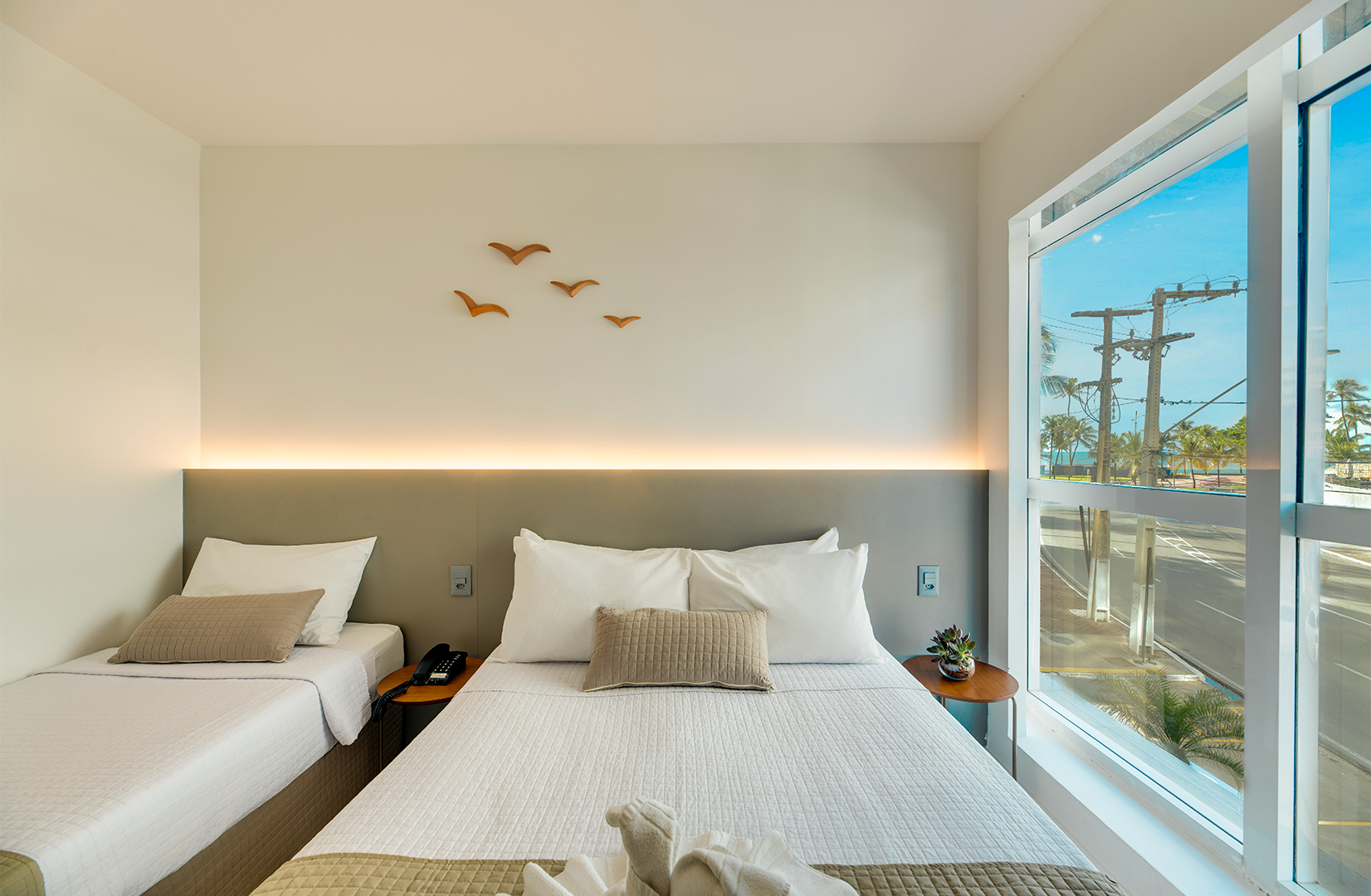 Quarto com três camas brancas e parede com painéis de madeira e iluminação zenital.