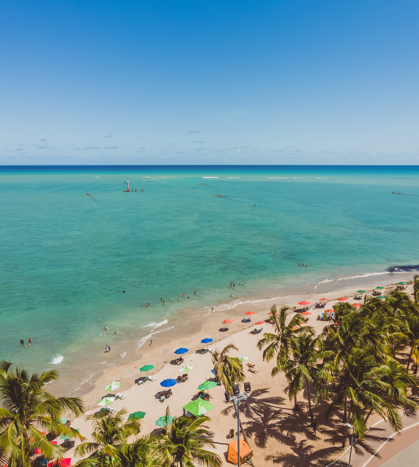 Oceano azul-turquesa com barcos navegando perto de uma praia com pessoas e edifícios em um dia ensolarado.