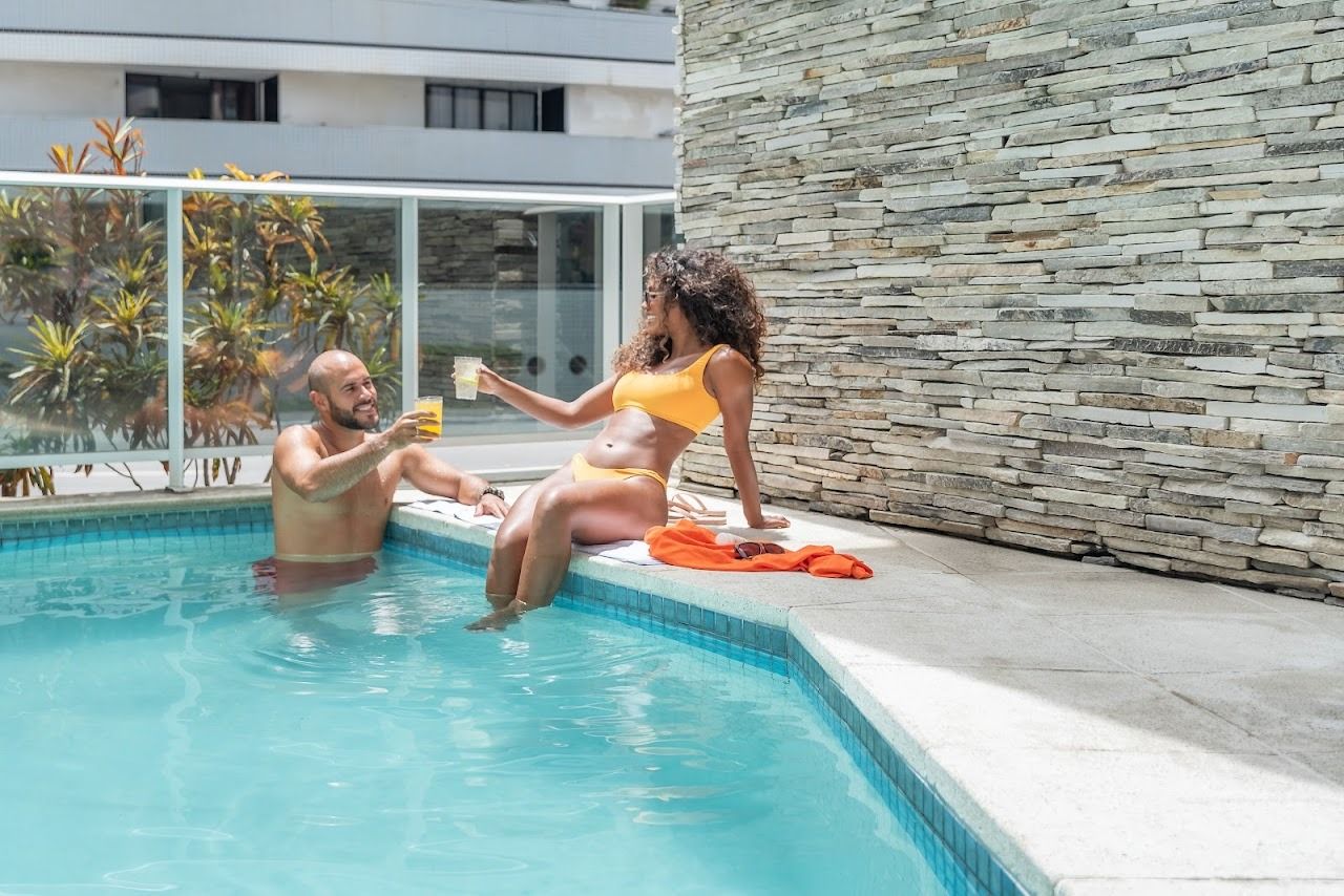 Um homem e uma mulher brindam com bebidas à beira da piscina. A mulher está sentada à beira da piscina, o homem na água.