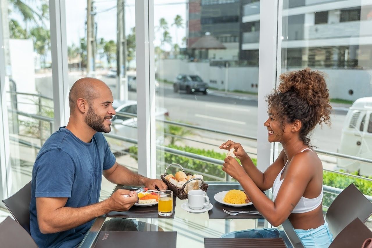 Homem e mulher sorrindo enquanto comem em uma mesa em frente a uma grande janela.