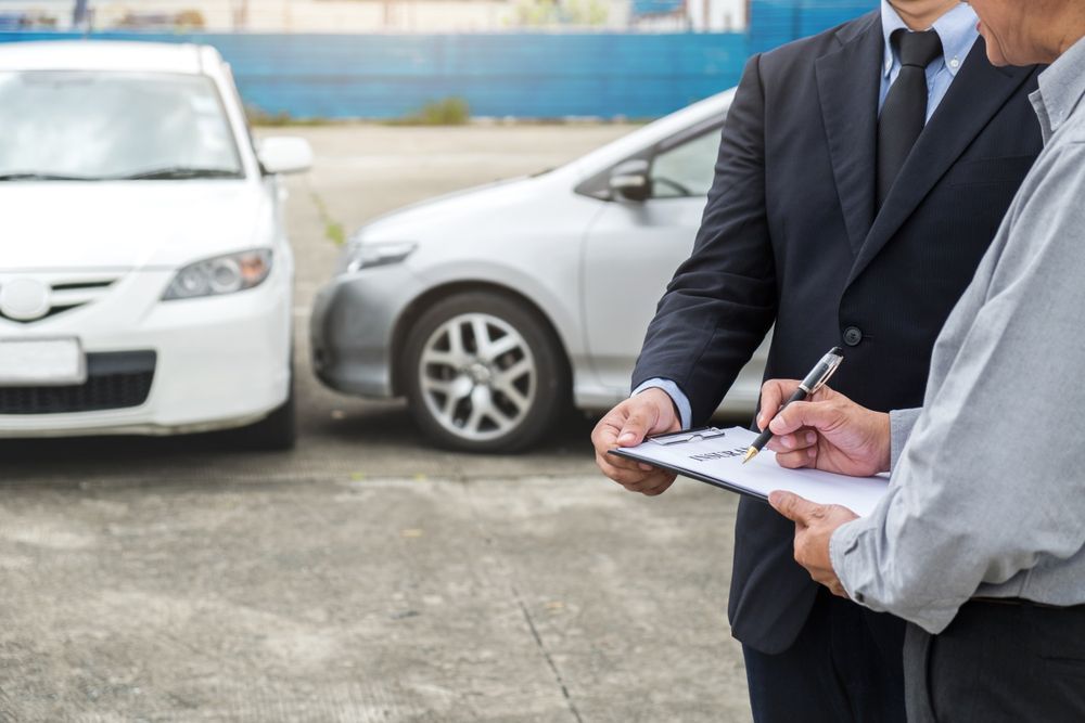 A Man in a Suit is Holding a Clipboard in Front of Two Cars — Blackwall Tyre Service & Mechanical Repairs In Blackwall, NSW
