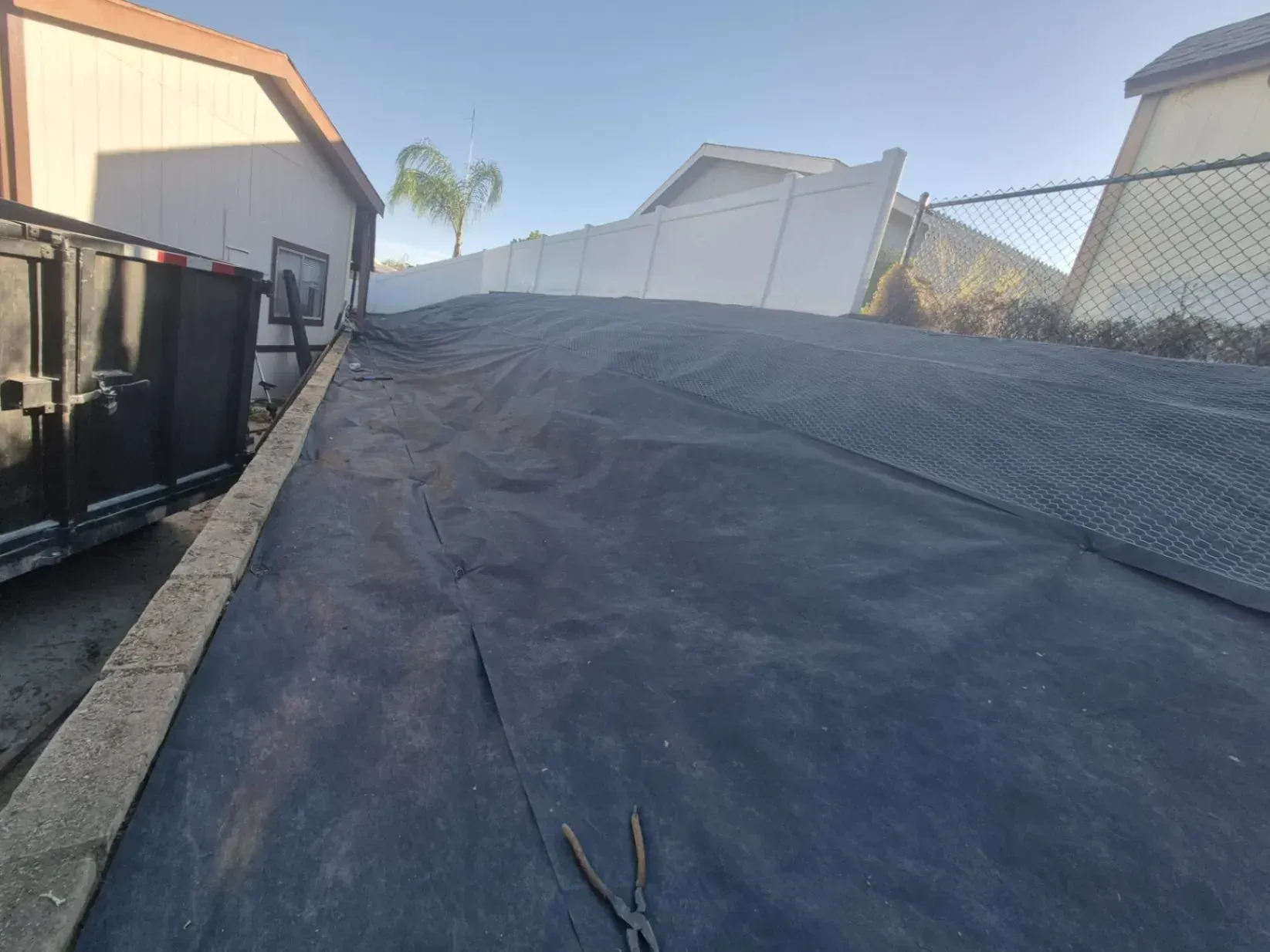 Black landscape fabric covers a sloped area, leading toward a white fence. A dumpster is on the left.