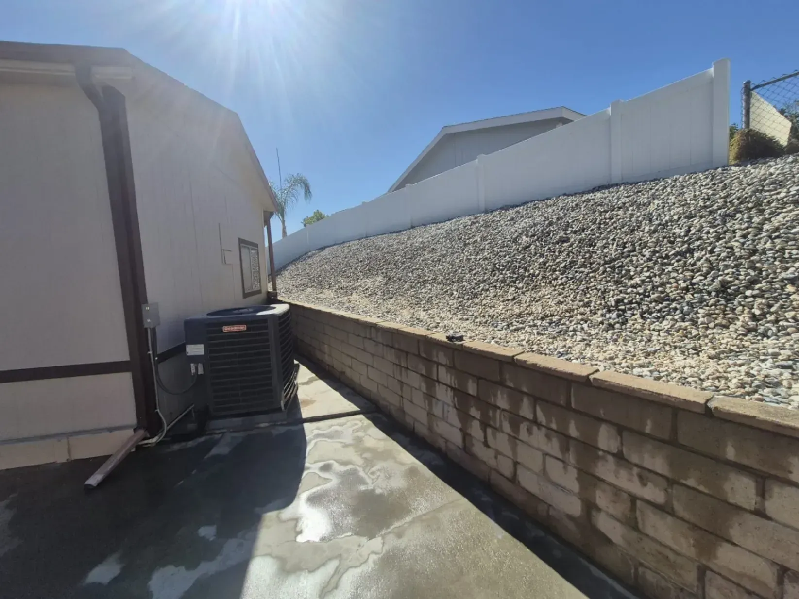 Side view of a house next to a retaining wall and gravel hillside, under a bright, sunny sky.