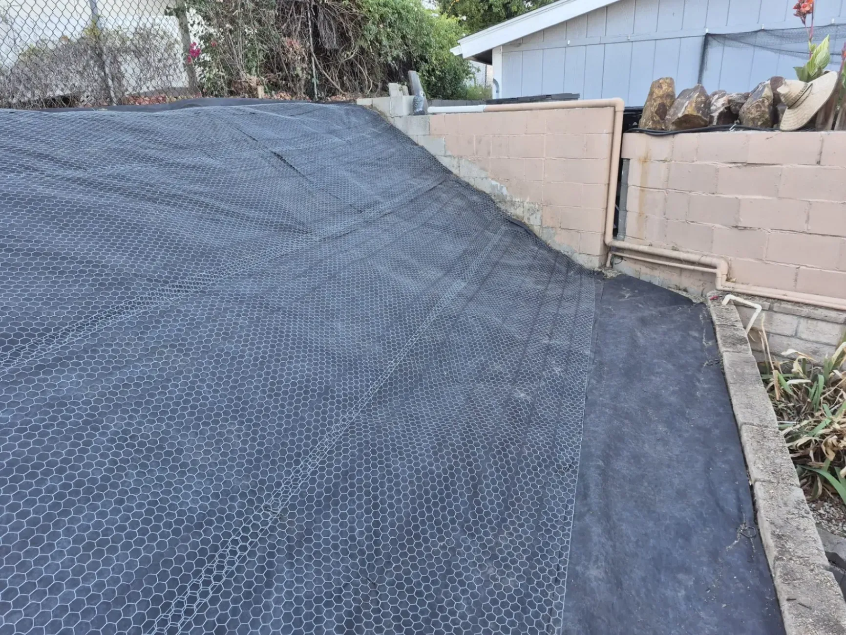 Black mesh and fabric cover a sloped surface adjacent to a tan concrete wall.