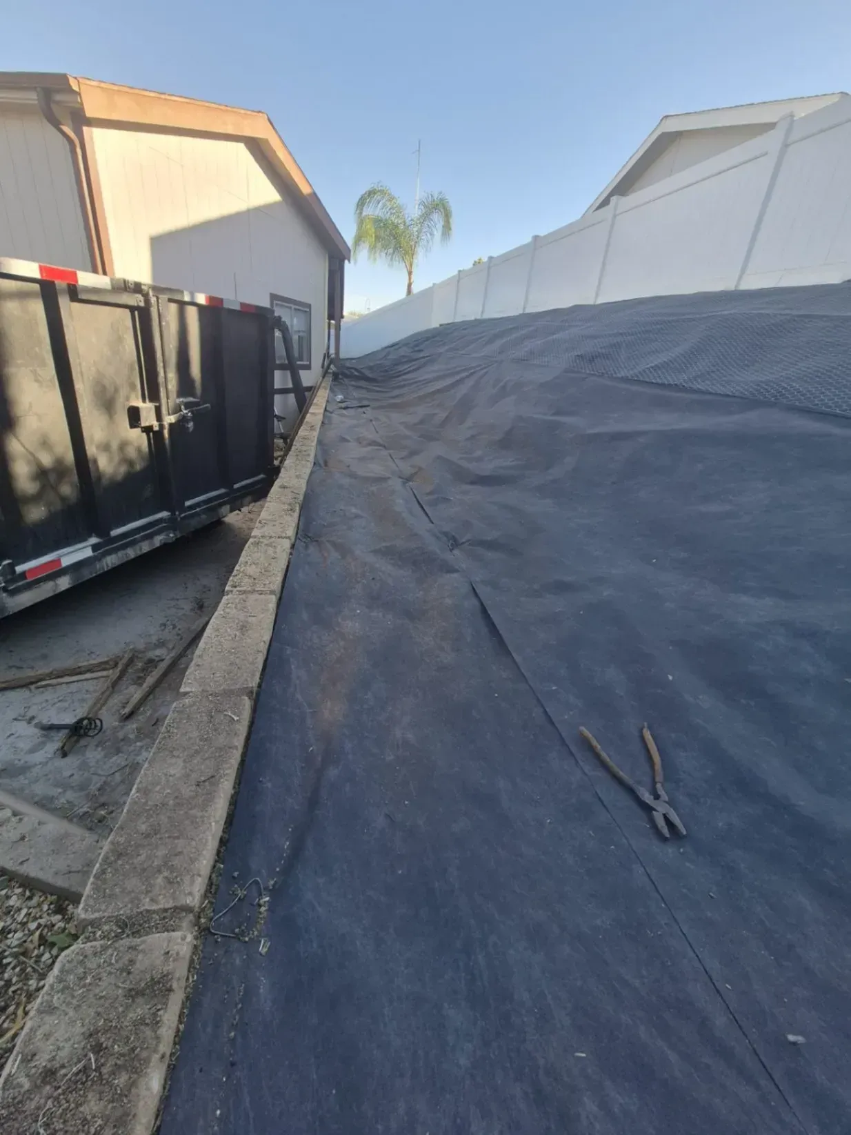 Black landscaping fabric covers a sloped yard, next to a retaining wall and buildings.