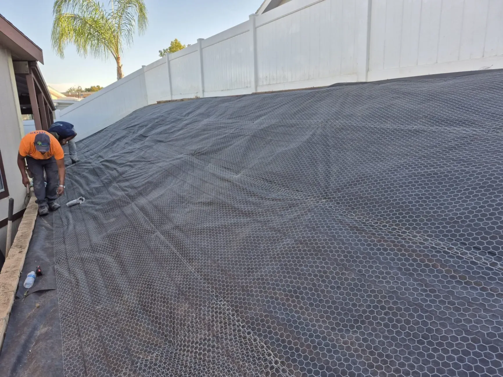 Two workers installing erosion control mesh on a sloped area next to a white fence.