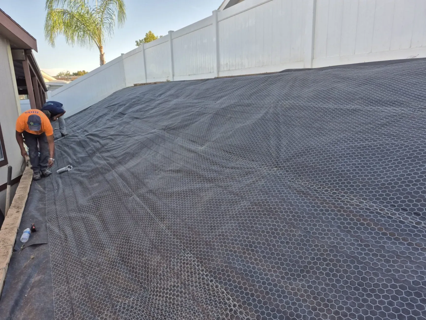 Two workers installing black mesh on a sloped area next to a white fence.