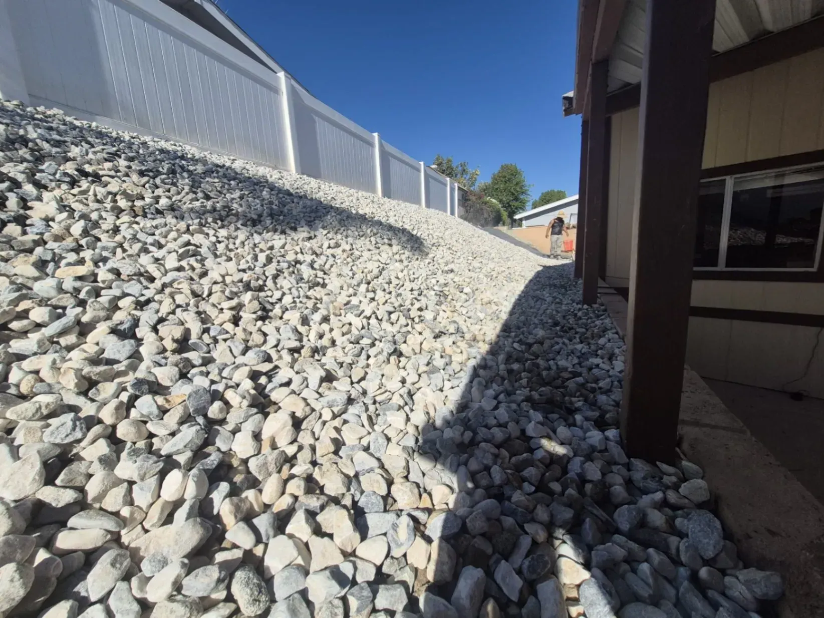 Rock-covered hillside next to a white fence and building with a brown support beam. Sunny, clear blue sky.