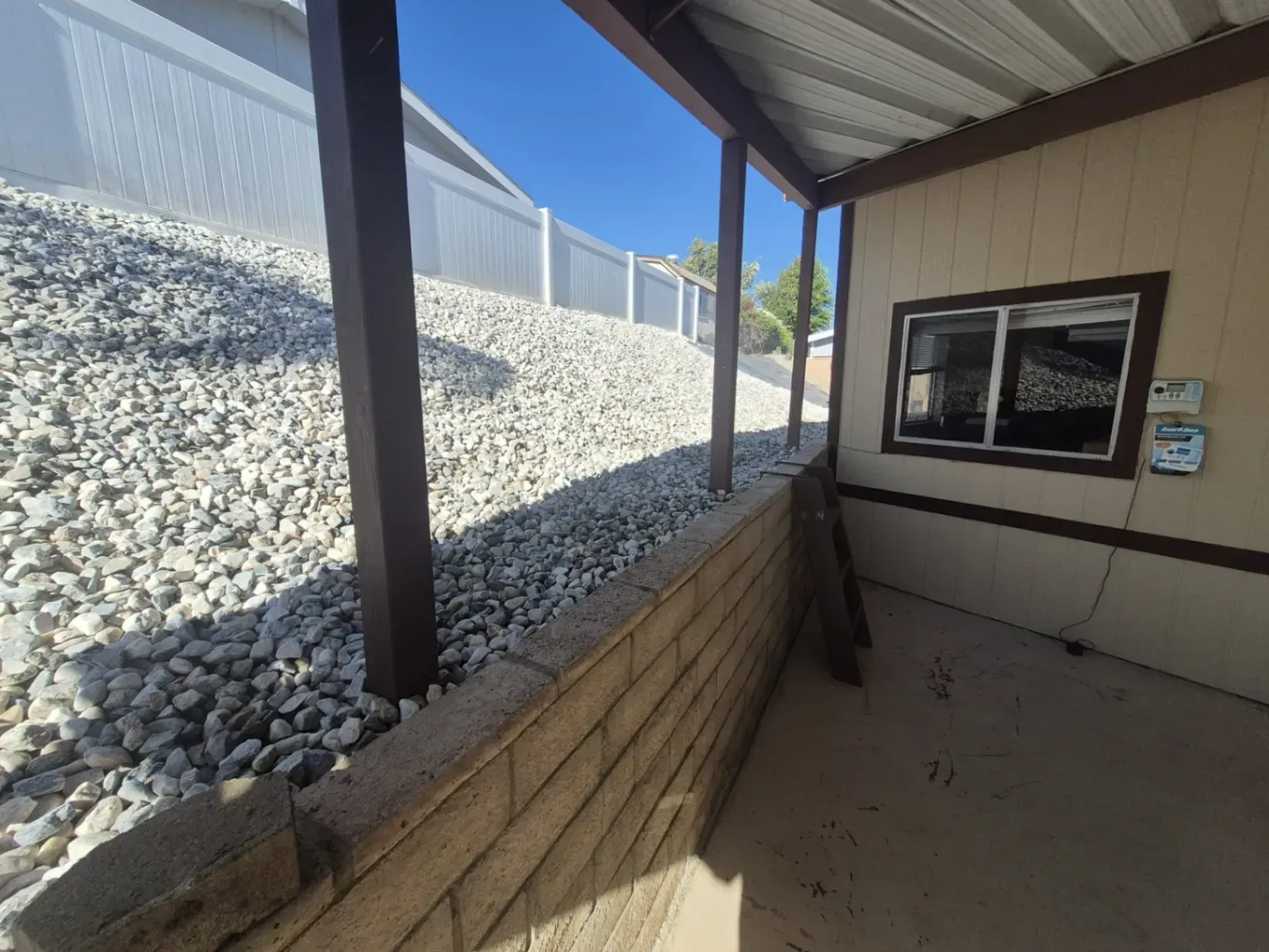Covered patio with a retaining wall next to a hillside covered in rocks. White fence and blue sky visible.