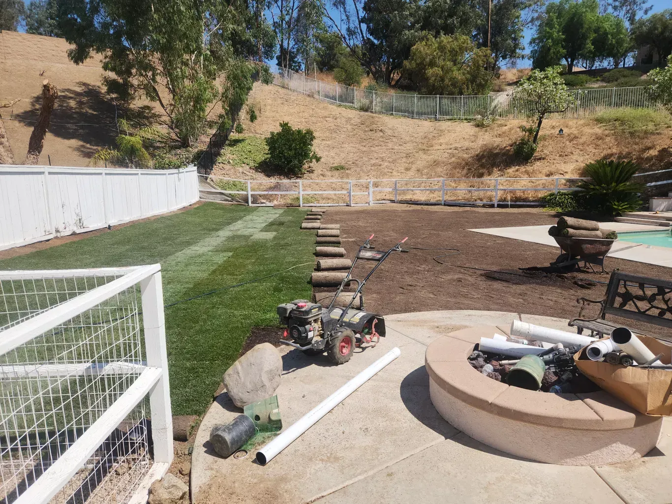 Backyard with freshly laid grass, fire pit, and fence; slope in the background.